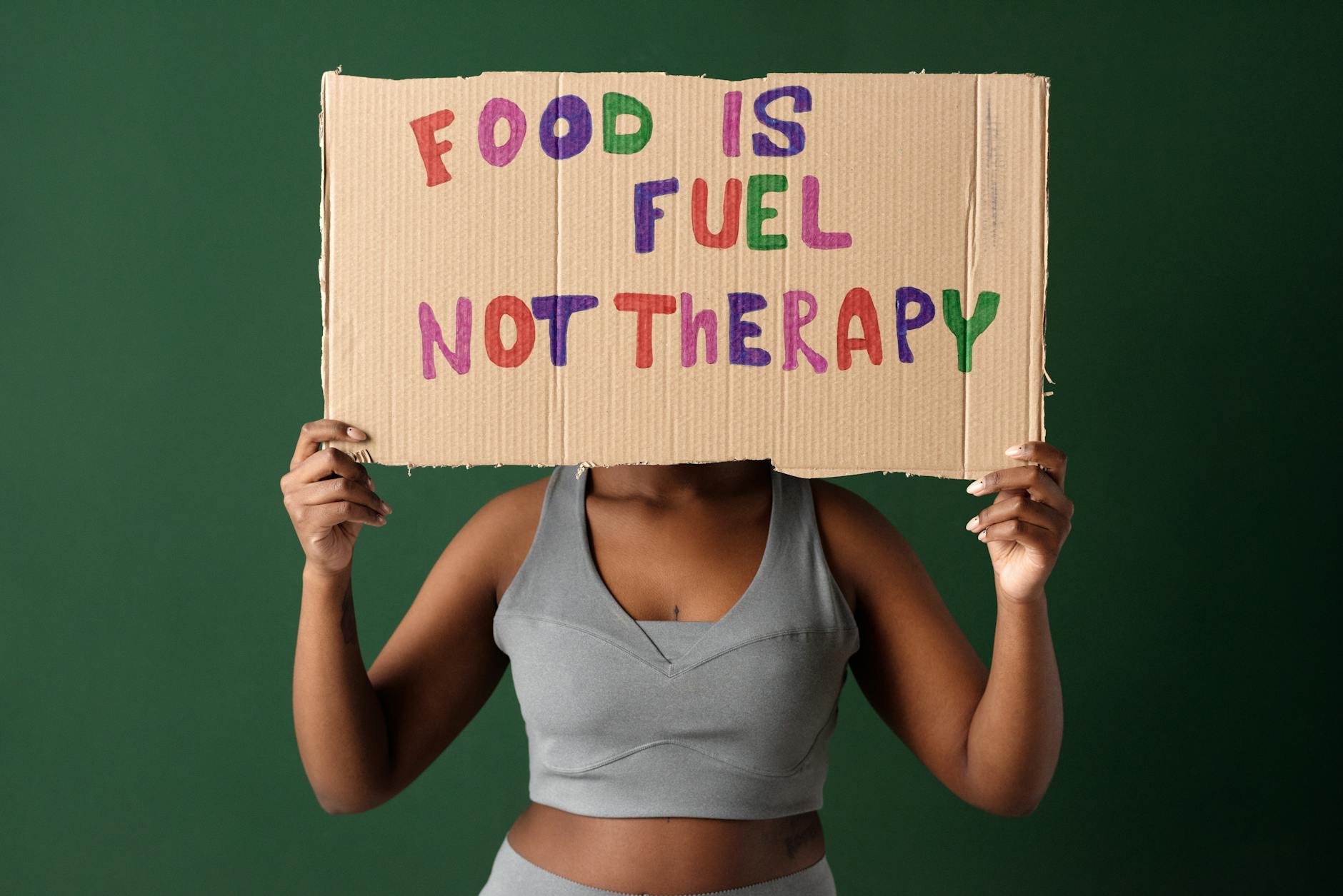 A woman holds a sign saying 'Food Is Fuel Not Therapy' against a green background.