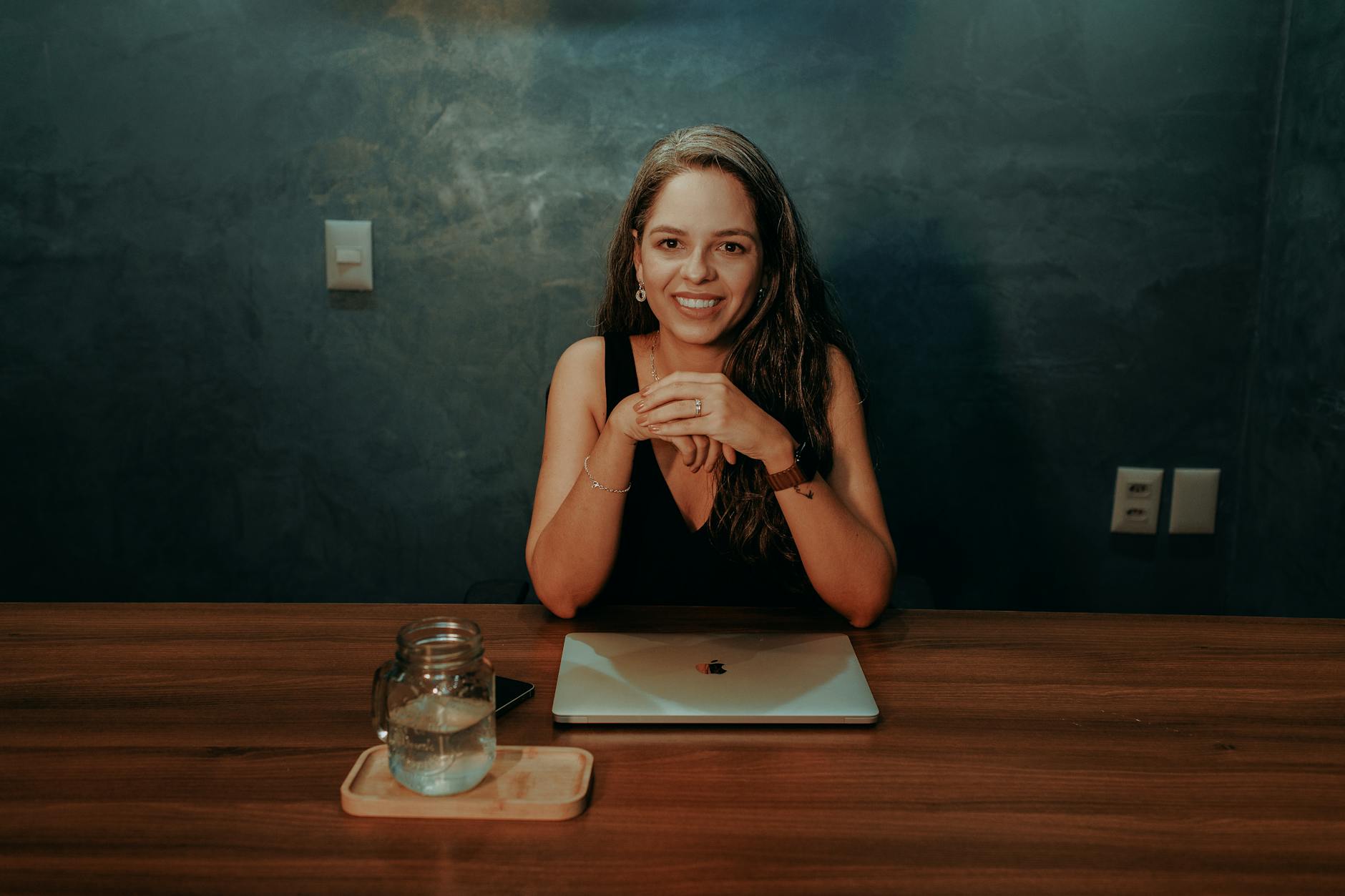 A woman smiling at a wooden desk with a laptop and jar, creating a cozy workspace vibe.