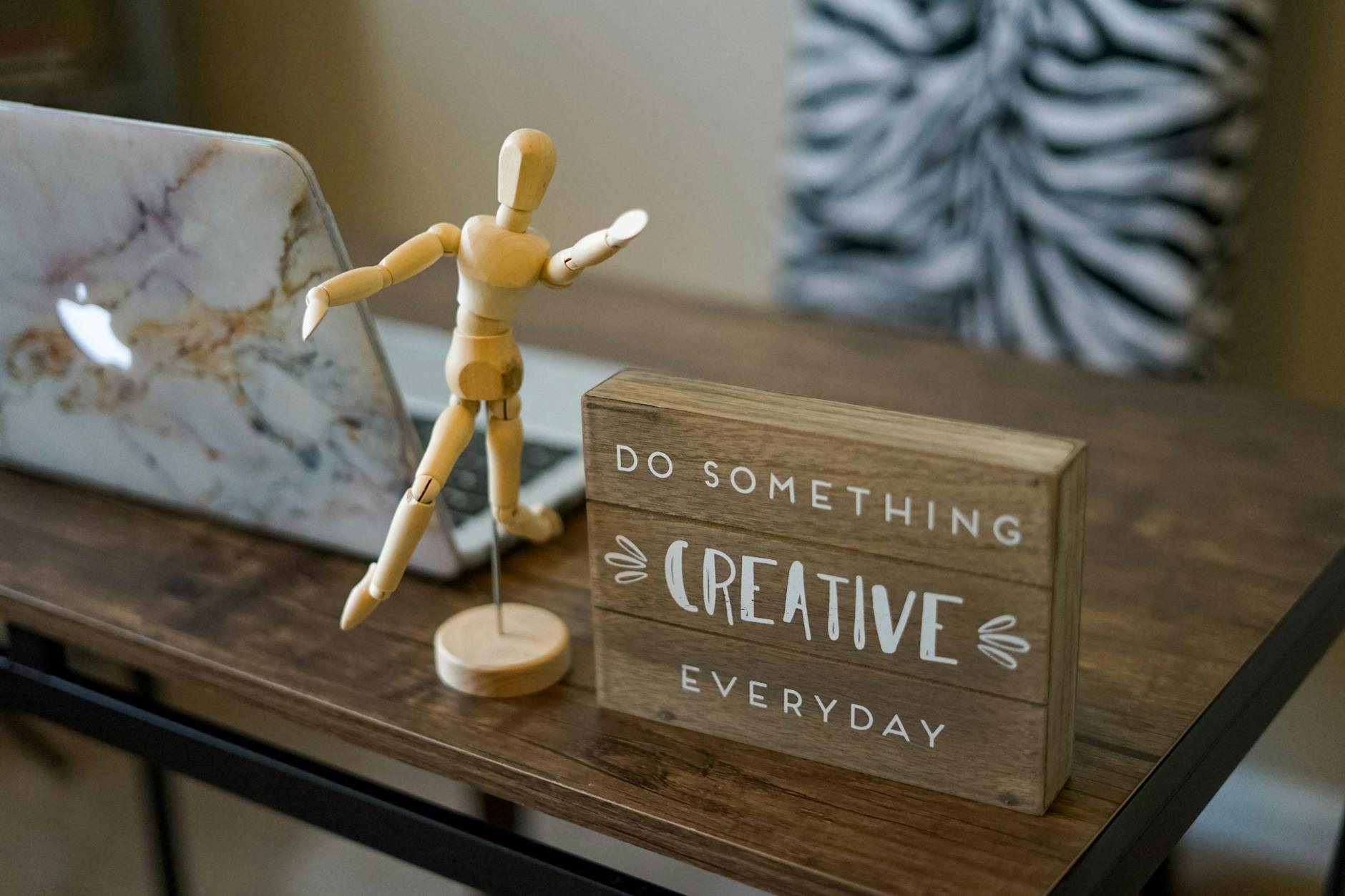 Wooden mannequin and inspirational sign on a desk beside a laptop, promoting creativity.