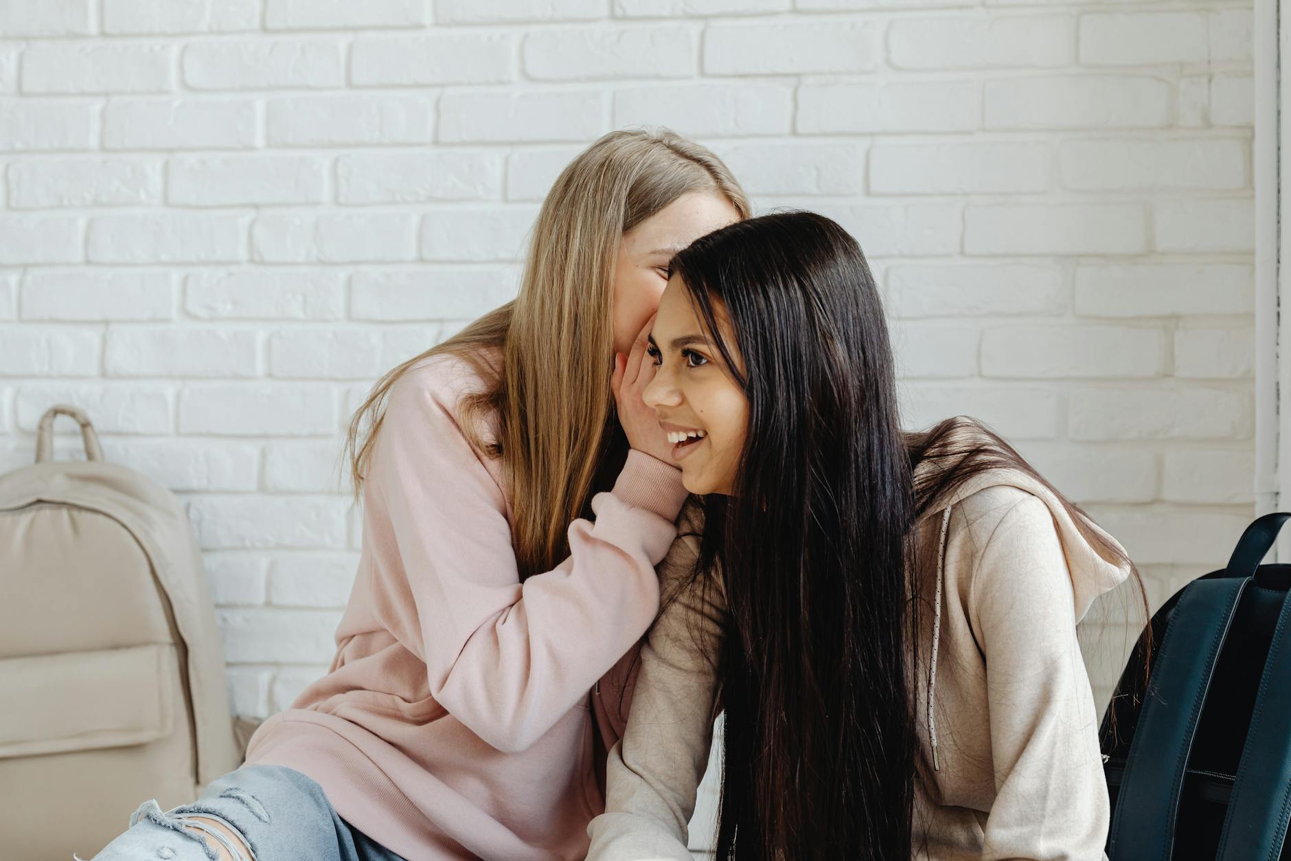 Two women in casual attire whisper and laugh against a white brick wall.