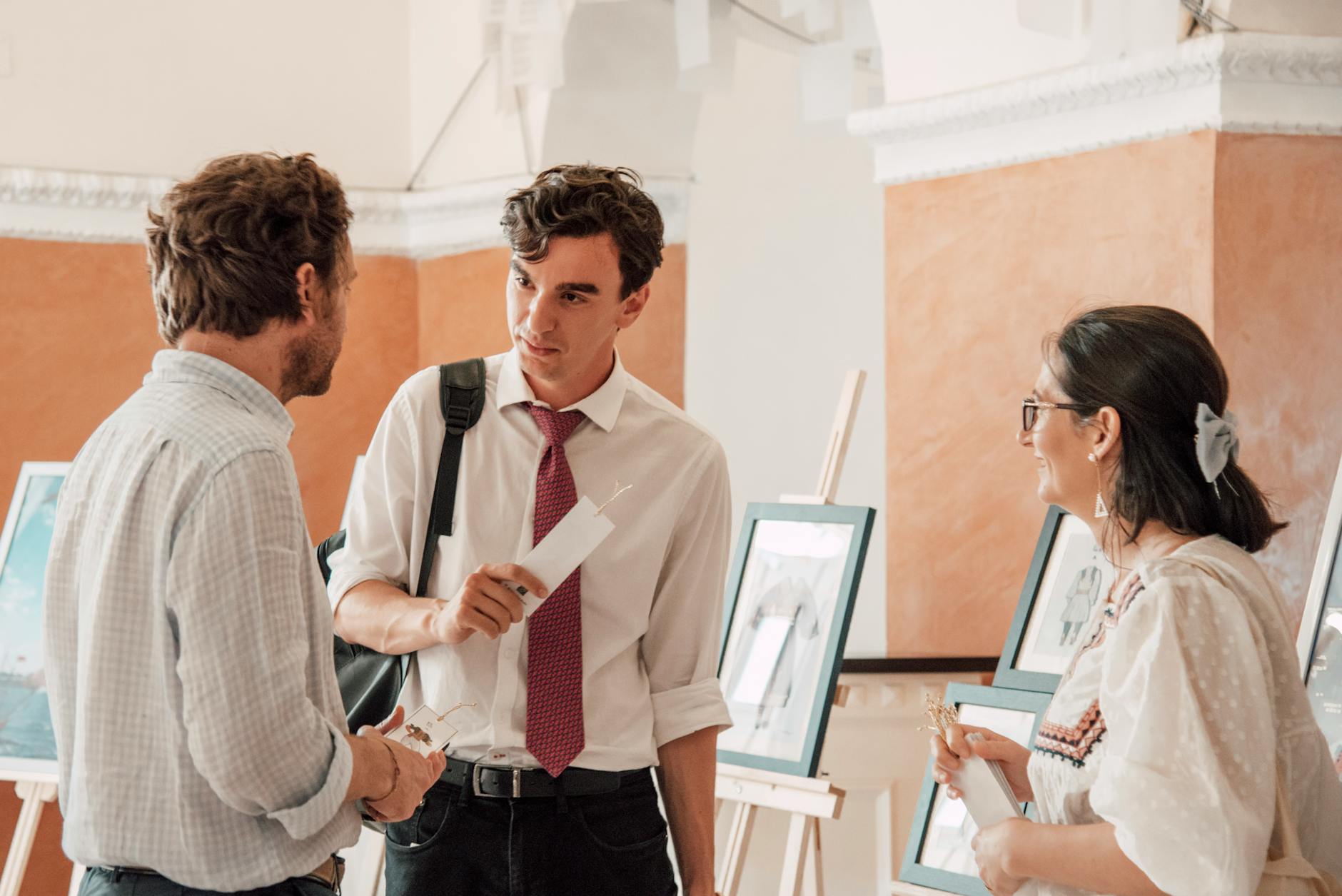 Three adults engaged in conversation at an art gallery exhibition with paintings on display.