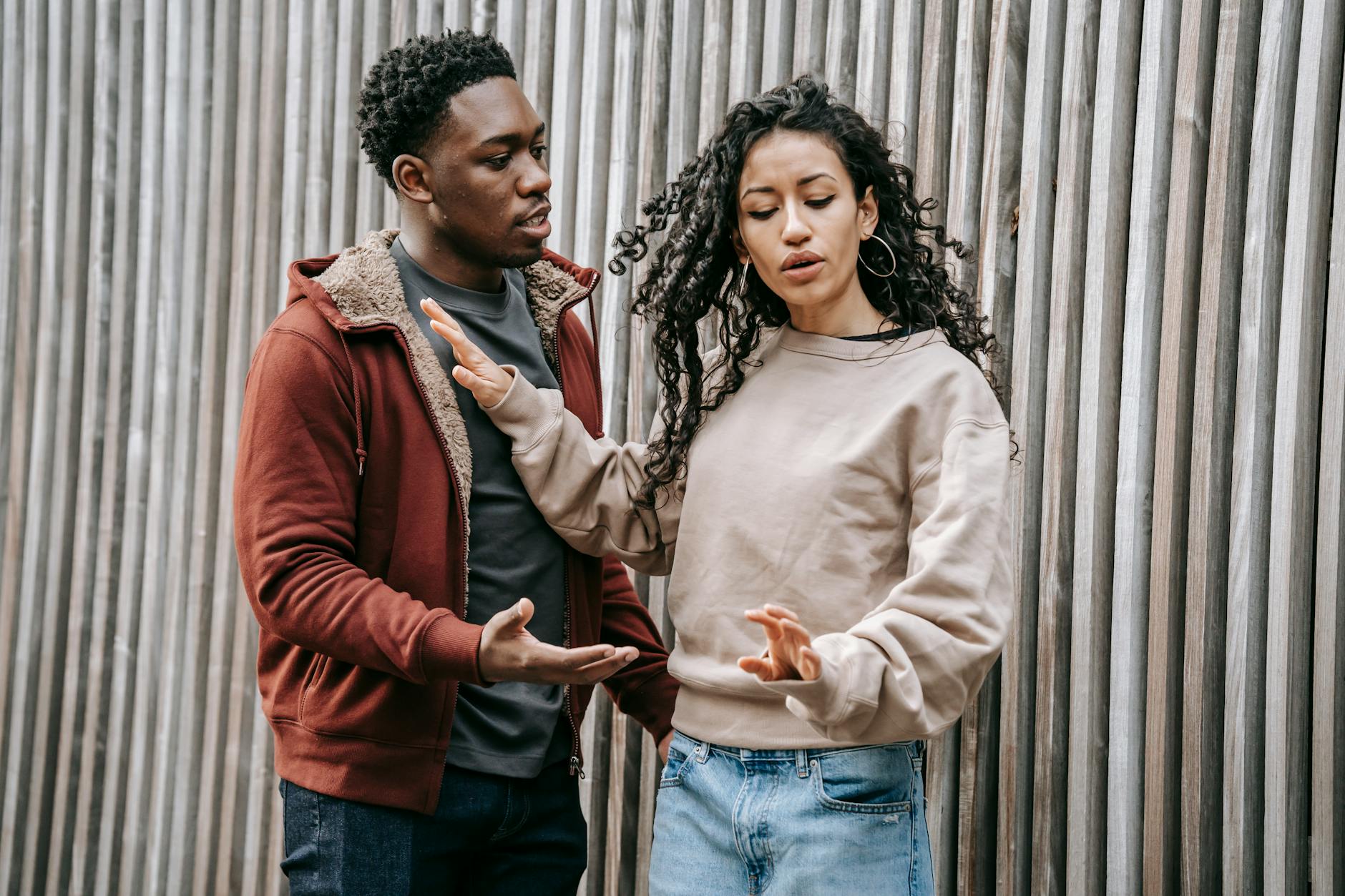 Annoyed ethnic girlfriend with curly hair pushing away concerned African American boyfriend while having conflict on street near wooden wall during breakup