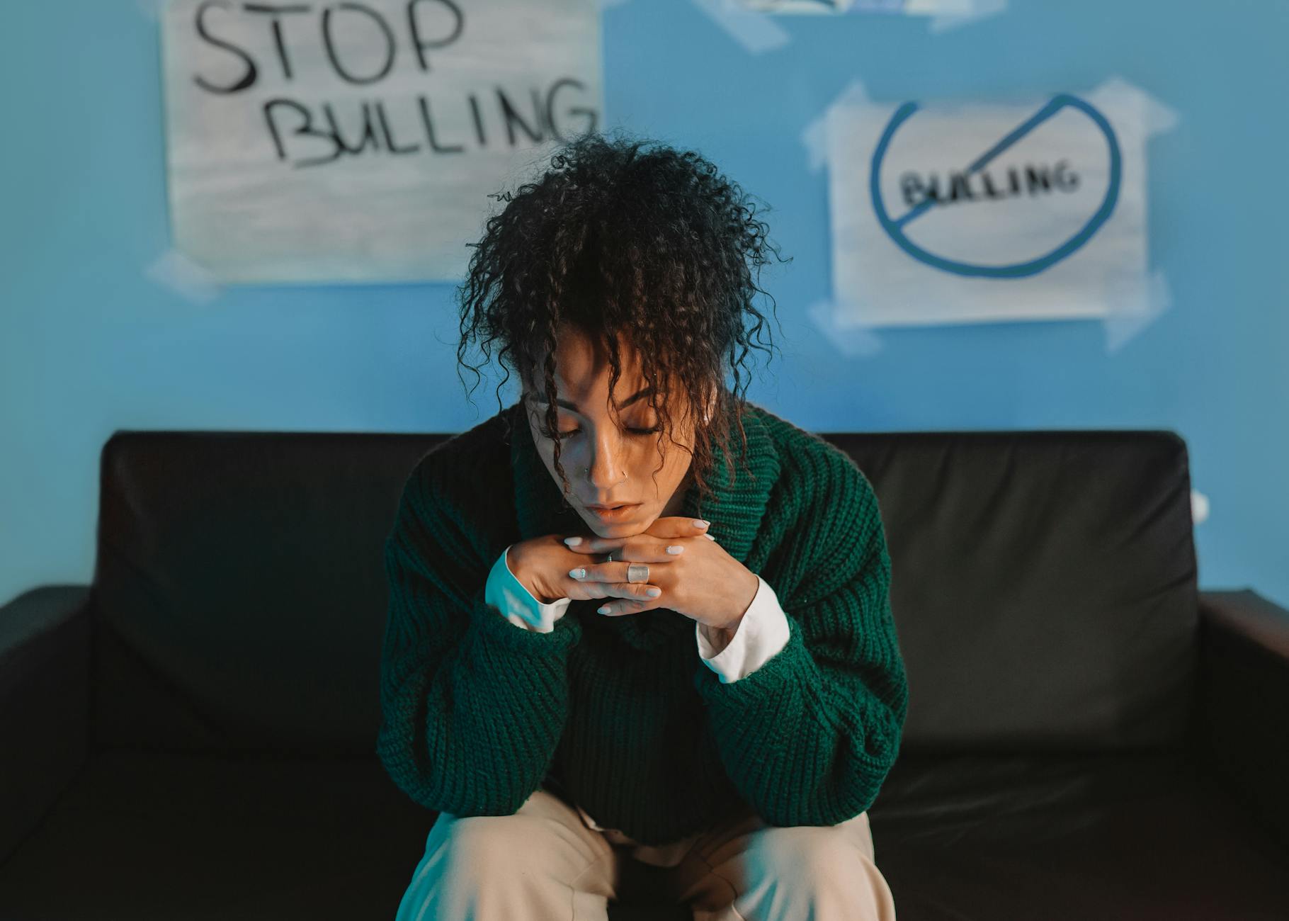 A woman sits sadly on a couch surrounded by anti-bullying posters in a reflective indoor setting.