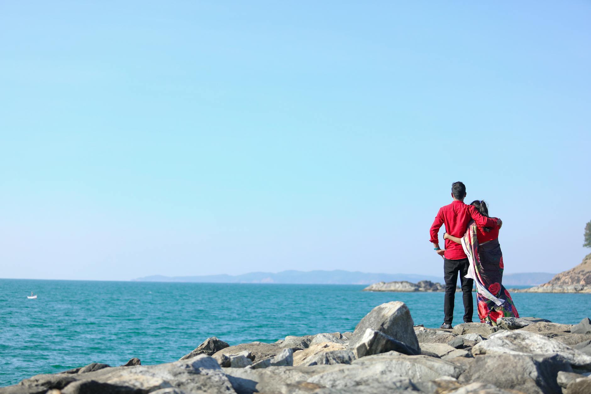 A couple standing on rocks embracing the scenic ocean view under a clear sky.