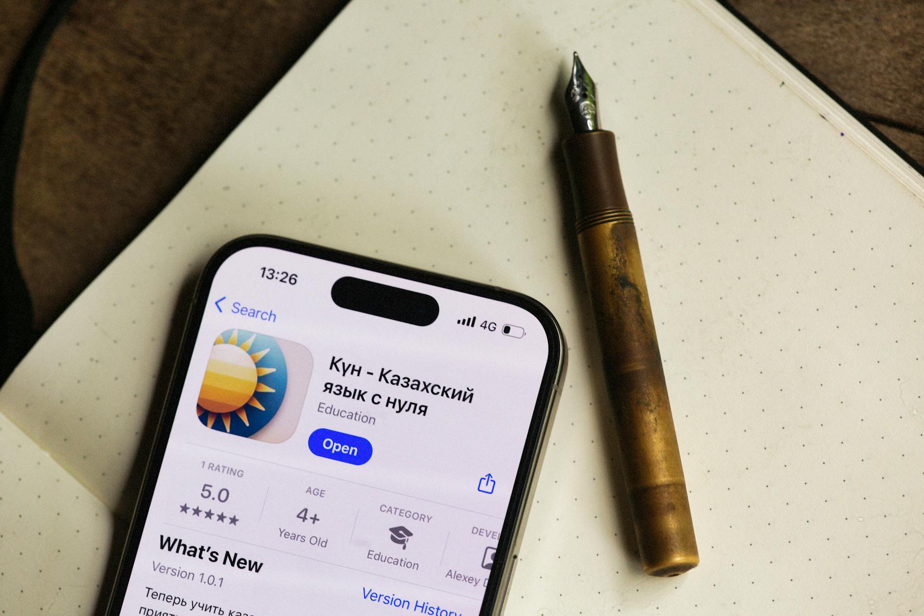 Smartphone displaying Kazakh language app next to a fountain pen on a notebook.