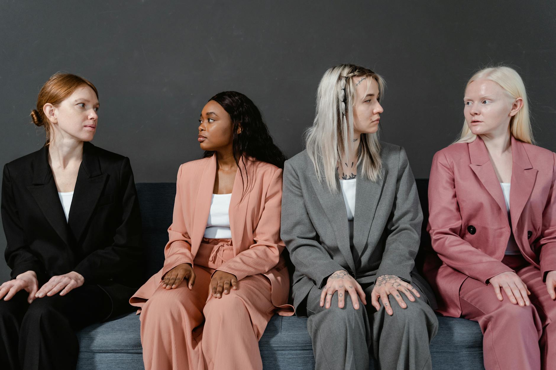 Four diverse women in formal attire sitting on a sofa, engaged in thoughtful interaction.