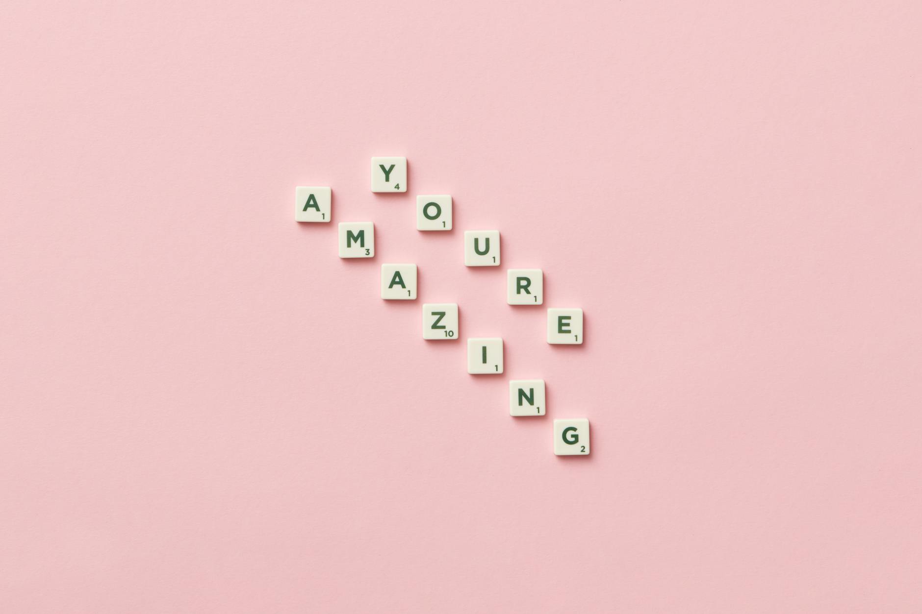 Inspirational quote 'You Are Amazing' spelled with Scrabble tiles on a pastel pink background.