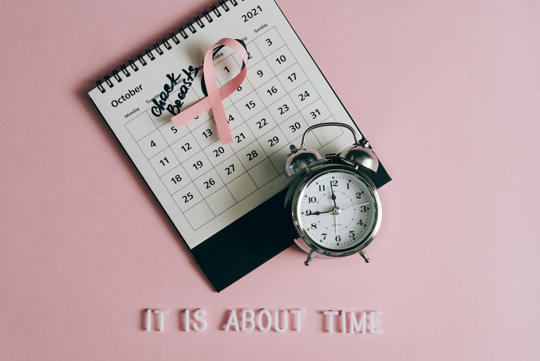 Calendar with pink ribbon, an alarm clock, and the text "Check breasts" signifies Breast Cancer Awareness.