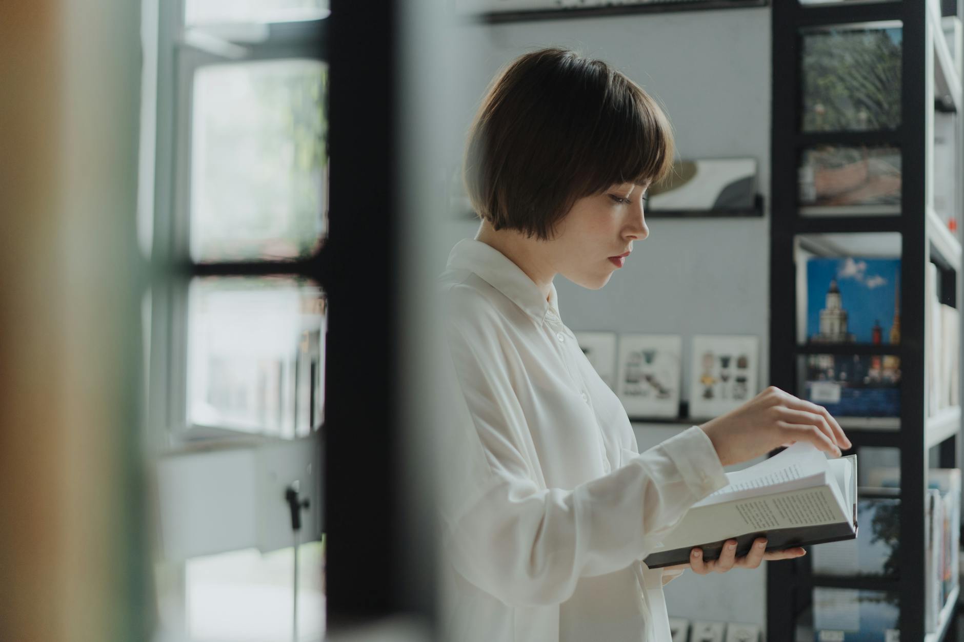 A young woman in a white shirt reads a book in a modern library setting, surrounded by bookshelves.