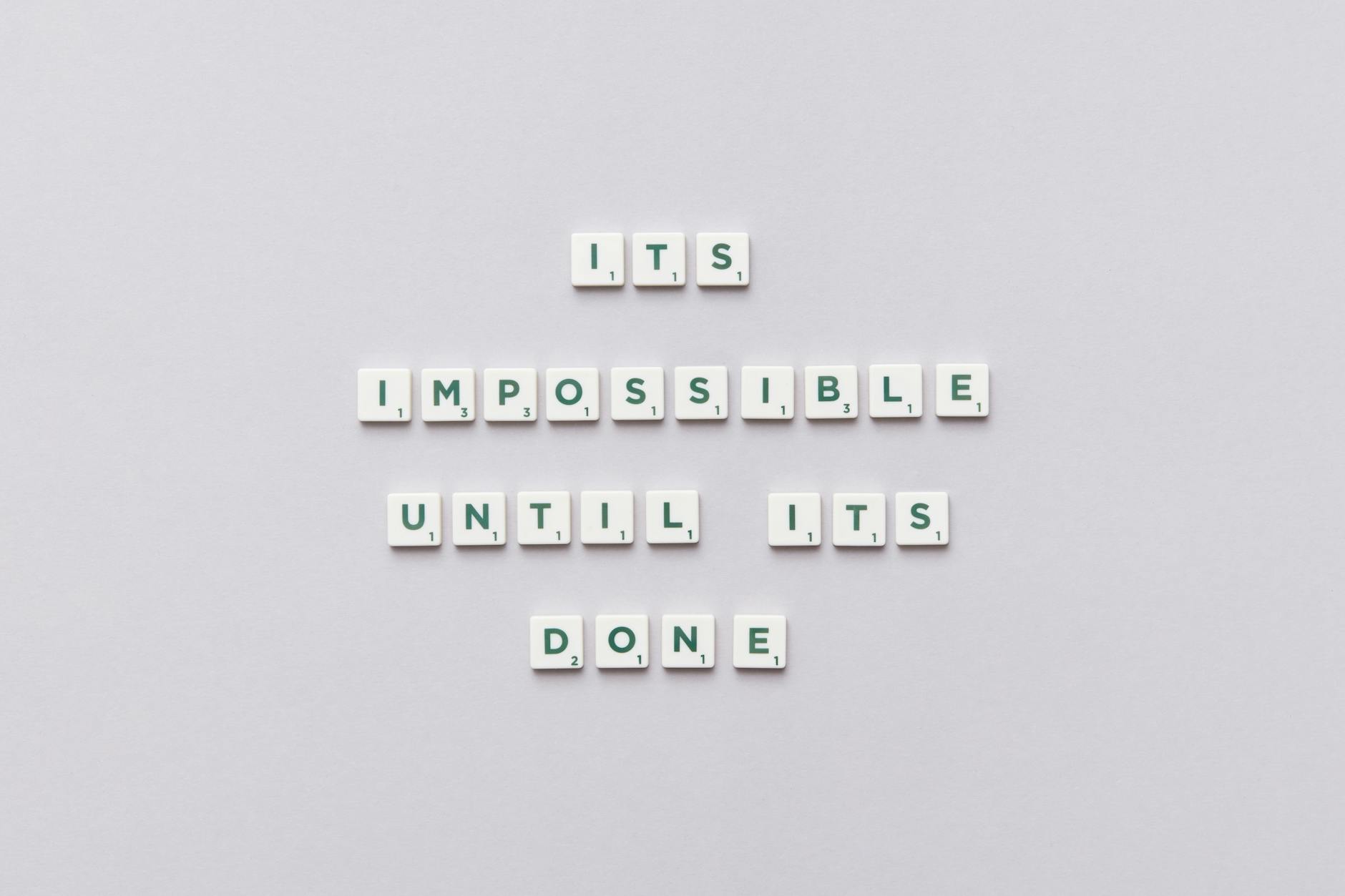 Motivational phrase 'It's impossible until it's done' arranged with Scrabble tiles on a gray background.