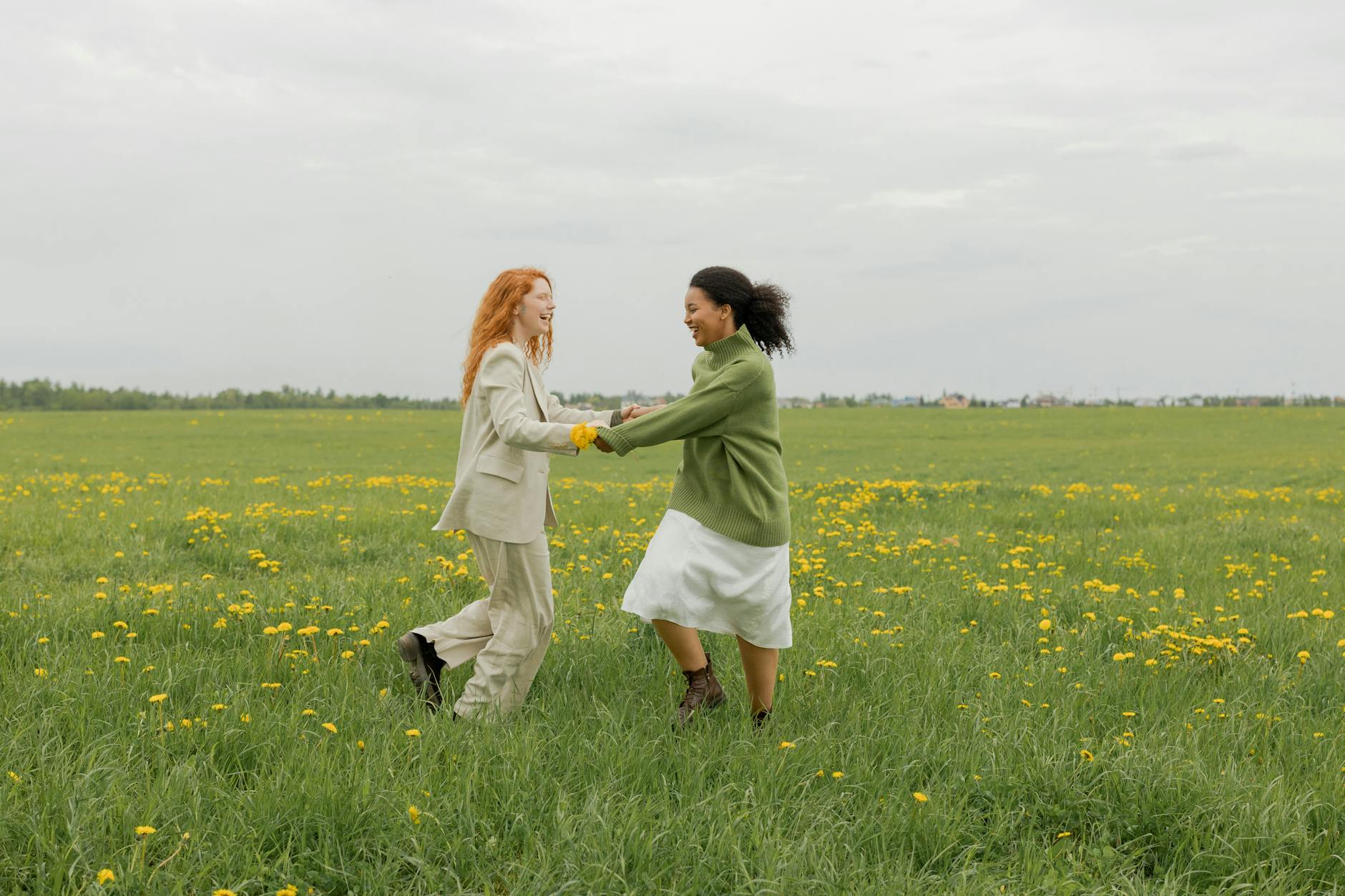Two women joyfully dance in a dandelion-filled field on a sunny day, embodying happiness and freedom.