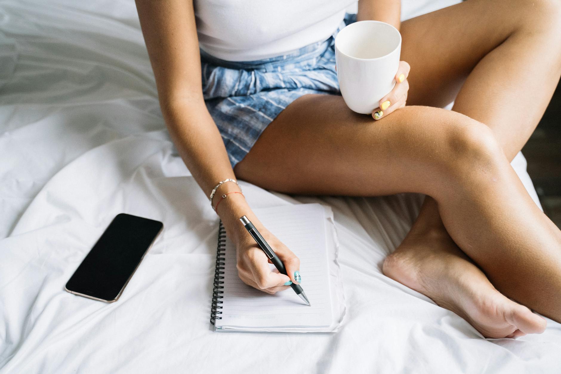 A woman holding a coffee cup while writing in a notebook, resting in bed.