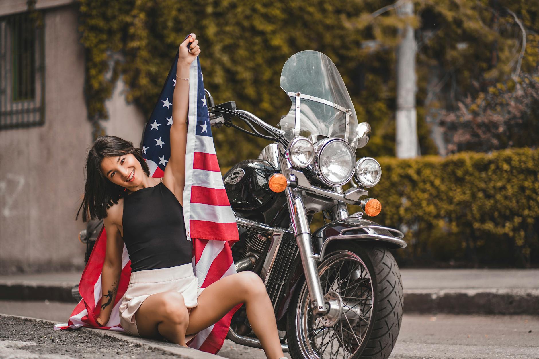 Young woman smiling while holding an American flag and sitting by a vintage motorcycle outdoors.